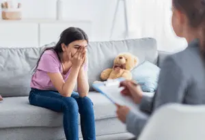 a teen buries their head in their hands next to a teddy bear on a couch during a panic disorder treatment program