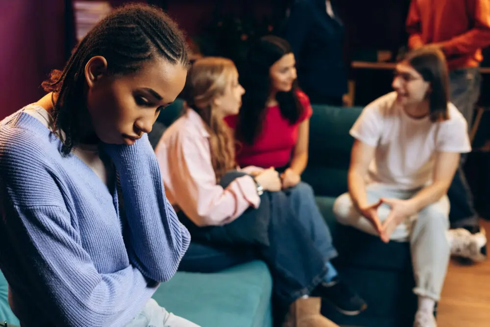 Teenager sitting alone while a group of friends talks in the background.