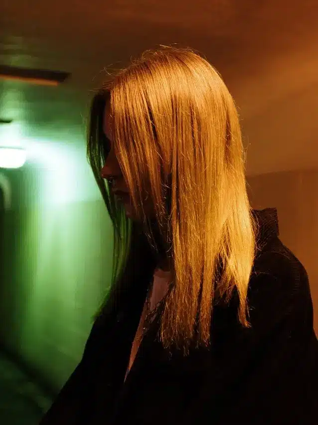 Knowing the Difference Between Normal Teenage Behavior and Possible Mental Health Disorders; blonde teen in a black hoodie stands in a green dimly lit tunnel with her hair covering her face